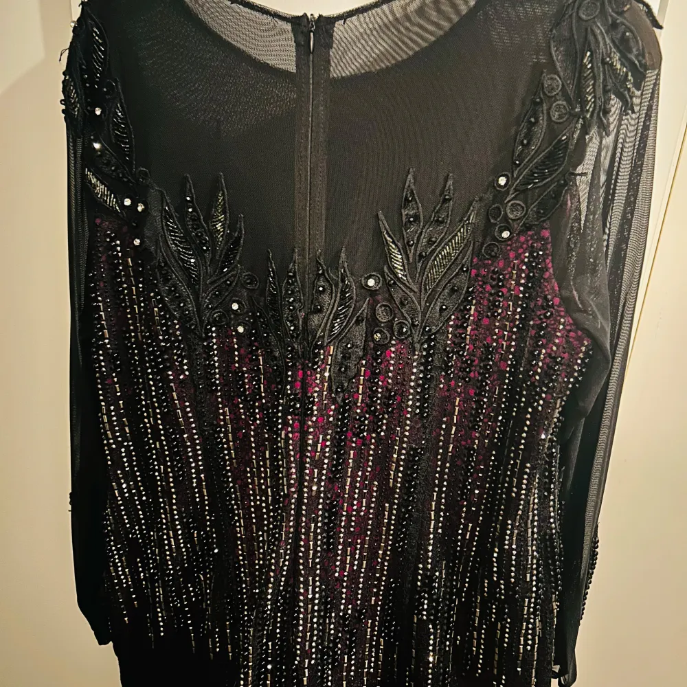 Elegant svart och lila långklänning med glittrande paljetter och detaljerad brodering. Klänningen har långa ärmar och en vacker fallande kjol med en hint av lila underton. Perfekt för en glamorös kväll.. Mekot.