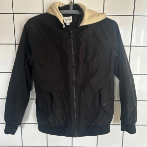 Svart bomberjacka från Kappahl - Snygg svart bomberjacka från Kappahl med beige huva. Jackan har dragkedja framtill och muddar vid ärmslut och nederkant. Perfekt för en cool och avslappnad stil.