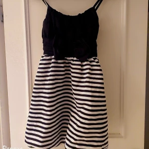 Randig klänning till flicka  från Lindex,storlek 152 - Söt klänning från Lindex med svart överdel och svartvit randig kjol. Klänningen har smala axelband och en stor rosett i midjan som ger en extra touch. Perfekt för sommarens äventyr!