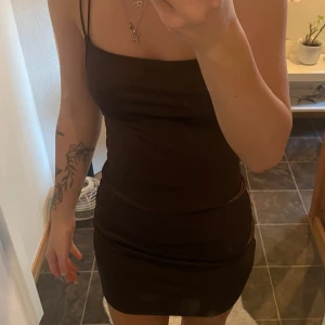 Brun tight klänning med smala axelband - Snygg brun klänning med smala axelband och korsade band i ryggen. Perfekt för en kväll ute. Den har en tight passform som framhäver figuren.