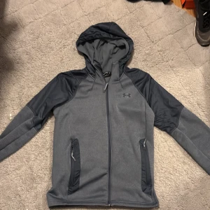 Grå hoodie från Under Armour - Säljer en grå hoodie från Under Armour med dragkedja och huva. Jackan har långa ärmar och två fickor med dragkedja framtill. Perfekt för en sportig look. Priset kan ändras via snabb affär