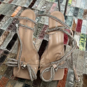 Beige sandaler med glittriga detaljer - Eleganta beige klackar med glittriga snörningar och en dekorativ rosett framtill. Sandalerna har en öppen tå och en transparent klack som ger en modern touch. Perfekta för att lägga till lite glamour till din outfit. Kanske använda 1 gång 