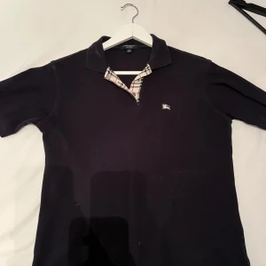 Svart pikétröja från Burberry - Snygg svart pikétröja från Burberry med klassisk krage och rutigt mönster på insidan av kragen. Tillverkad i 100% bomull för en bekväm passform. Kom med prisförslag 