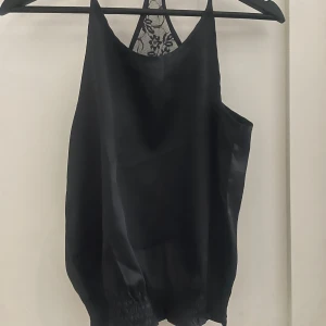 Svart linne med spetsdetaljer från XOXO & friends - Snygg svart linne från XOXO & friends med spetsdetaljer i ryggen. Perfekt för en stilren look. Tillverkad i ett glansigt material som ger en lyxig känsla.