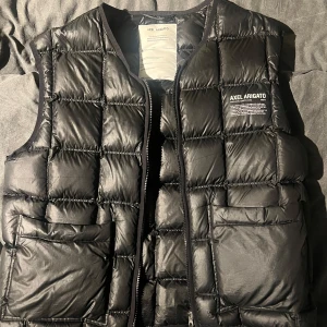 Axel Arigato Binx Puffer West - Snygg svart dunväst från Axel Arigato med quiltad design och dragkedja framtill. Västen har praktiska fickor och är tillverkad av återvunnen polyester och dun. Perfekt för att hålla värmen med stil.