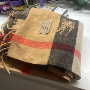 Burberry scarf - Snygg och klassisk rutig halsduk från Burberry i beige, svart och röd. Tillverkad i mjukt material med broderad logotyp och etikett. Perfekt för att hålla dig varm och stilren under kyliga dagar.