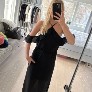 Svart långklänning med volangdetaljer - Snygg svart långklänning med volangdetaljer och en elegant oneshoulder-design. Perfekt för en stilren look med en smickrande passform. Klänningen har en elastisk midja och tunna axelband som korsas i ryggen. Storlek 34, XS. Märket är Divided H&M