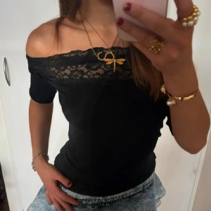 Svart offshoulder topp med spetsdetalj - Snygg svart offshoulder topp med spetsdetalj vid halsen. Perfekt för en stilren look. Toppen har korta ärmar och en figurnära passform. Skriv för bilder då denna är lånad!