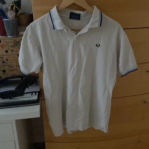 Vit pikétröja från Fred Perry - Säljer en klassisk vit pikétröja från Fred Perry med blå detaljer på krage och ärmslut. Tröjan har en broderad logotyp på bröstet och är tillverkad i 100% bomull. Perfekt för en stilren look. Ass skön i storlek medium men den är liten i storlek så den passar mig som har storlek s