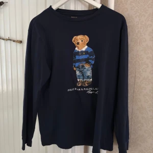 Ralph Lauren Mörkblå långärmad tröja med Polo Bear tryck - Säljer en mörkblå långärmad tröja från Ralph Lauren med ett polo björntryck på framsidan. Tröjan har en klassisk rund hals och är perfekt för en avslappnad stil. Den är i storlek 170/Barn XL.