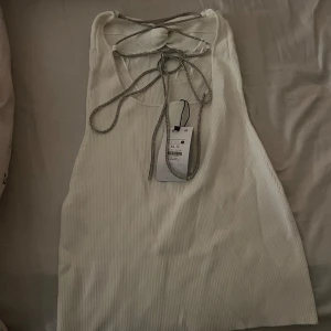 Vit ribbad klänning från Bershka - Säljer en stilren vit ribbad klänning från Bershka. Klänning har en snygg knytning i nacken och är ärmlös, perfekt för varma dagar. Den är i storlek XL men är lite liten i storleken 