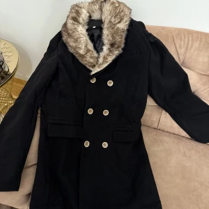 Svart kappa med pälsdetalj - Elegant svart kappa med dubbelknäppning och en pälsdetalj runt kragen. Perfekt för att hålla sig varm och stilren under kyliga dagar. Kappan har långa ärmar och en klassisk design som passar till många tillfällen.