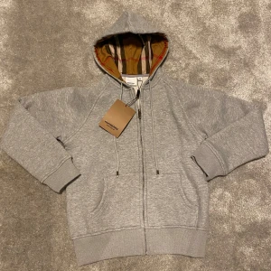 Grå hoodie från Burberry - Säljer en stilren grå hoodie från Burberry med klassiskt rutigt mönster i huvan. Tröjan har dragkedja framtill och ribbade muddar vid ärmslut och nederkant. Perfekt för en avslappnad och trendig look.