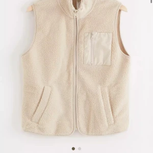  fleeceväst från Lindex - Säljer en mysig beige fleeceväst från Lindex med dragkedja och praktisk bröstficka. Perfekt för lager-på-lager under kyliga dagar. Västen har en enkel och stilren design med två sidofickor.
