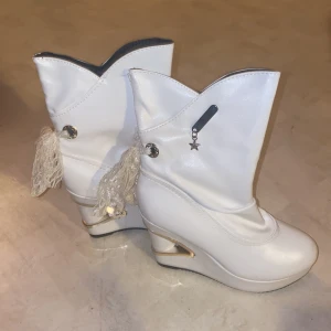 Vita boots med kilklack och stjärndetalj - Unika vita boots med kilklack och dekorativa tofsar på sidan. Skorna har en stjärndetalj och en elegant design som sticker ut. Perfekta för att ge en extra touch till din outfit.