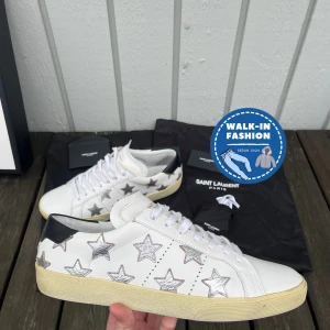 Vita sneakers med stjärnor från Saint Laurent - Snygga vita sneakers från Saint Laurent med silverfärgade stjärnor och svart häl. Skorna har klassisk snörning och en stilren design som passar perfekt för en trendig look.