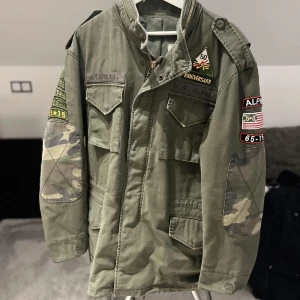 Grön fieldjacka med patchar - Säljer en grön militärinspirerad jacka från Alpha Industries. Jackan har flera patchar och detaljer som ger en autentisk look. Den har långa ärmar med camouflage-mönstrade armbågslappar och praktiska fickor framtill. Perfekt för en fet och avslappnad stil.