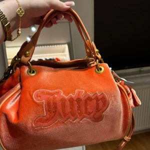 Y2k Orange sammetväska från Juicy Couture - Hellt ny och snygg orange y2k handväska i sammet från Juicy Couture med dekorativt Juicy-märke på framsidan. Väskan har guldfärgade detaljer och en söt rosett. Perfekt för att ge en färgklick till din outfit!