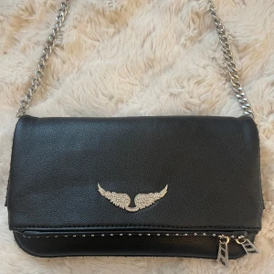 Svart axelväska med kedja och vingdetalj - Snygg svart axelväska i skinn från Zadig & Voltaire med en cool vingdetalj i silver på framsidan. Väskan har en kedjerem och små nitar längs kanten🤍ny pris är 4500, köpte den i Paris 😊