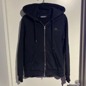 Svart zip hoodie från J.Lindeberg - Säljer en stilren svart zip hoodie från J.Lindeberg med dragkedja och justerbar huva. Tröjan har långa ärmar och praktiska fickor framtill. Perfekt för en avslappnad look. De är mycket bra skick förutom att några bokstäver på J.lindberg loggan släppt efter tvättning annars är det inga fel på tröjan. Ny pris 1199kr mitt 400kr