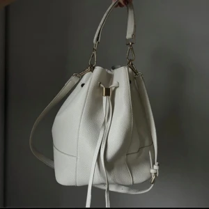 Vit axelväska - Snygg vit axelväska, väskan har en stilren design med knytband och metallspännen i guld. 