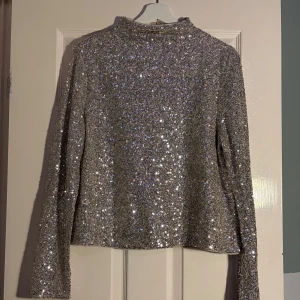 Glittrig silvertröja från H&M - Säljer en glittrig silvertröja från H&M. Tröjan har långa ärmar och en dragkedja i ryggen för enkel på- och avtagning. Perfekt för att lysa upp vilken outfit som helst med sitt glittrande paljettmönster. Passar utmärkt till festliga tillfällen! ✨