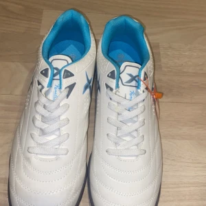 Vita fotbollsskor med blå detaljer (futsal) - Snygga vita fotbollsskor med blå insida och detaljer. Skorna har snörning och en slitstark sula med bra grepp för spel inom futsal. Perfekta för fotbollsträning eller match inomhus i FUTSAL.