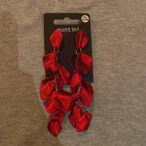Röda örhängen från Ur&penn  - Snygga röda örhängen från Montini med hängande blommor i satinliknande material. Perfekta för att ge en färgklick till din outfit. De har en elegant design med flera lager av röda blomblad som hänger i kedjor.