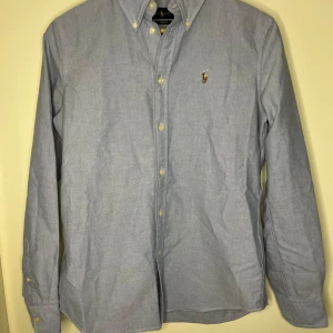 Blå skjorta från Ralph Lauren - Snygg blå skjorta från Ralph Lauren med klassisk button-down krage och broderad logga på bröstet. Skjortan har långa ärmar och knappar framtill. Perfekt för en stilren look.