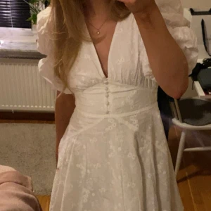 Adoore klänning - Säljer en söt vit Adoore- klänning med puffiga kortärmar och v-ringning. Klänningen har ett blommigt mönster och knappar framtill. Perfekt för studenten! 🌸 Perfekt skick och helt utan fläckar eller liknande! 💕Helt slutsåld på hemsidan och eftertraktad.