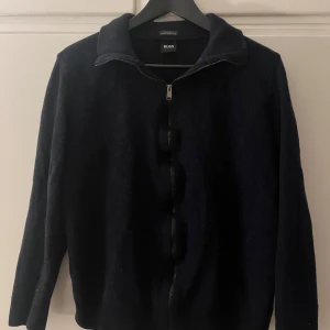 Svart tröja med dragkedja från Hugo Boss - Säljer en stilren svart tröja från Hugo Boss, tillverkad av finaste italienska garnet. Tröjan har en dragkedja framtill och långa ärmar, perfekt för en elegant look. Passformen är regular fit.