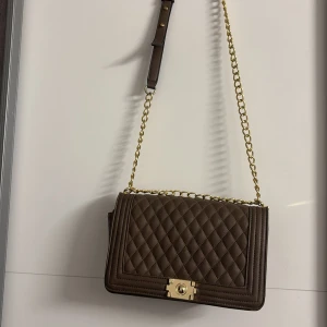 Brun quiltad axelväska med guldkedja - Snygg brun axelväska med quiltat mönster och guldkedja. Väskan har en elegant låsdetalj framtill och en praktisk dragkedja på sidan. Perfekt för att ge din outfit en stilfull touch.