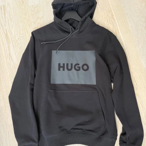 Svart hoodie från Hugo - Säljer en stilren svart hoodie från Hugo med en stor logga på bröstet. Tröjan har en praktisk känguruficka och justerbar huva med snören. Perfekt för en avslappnad look.