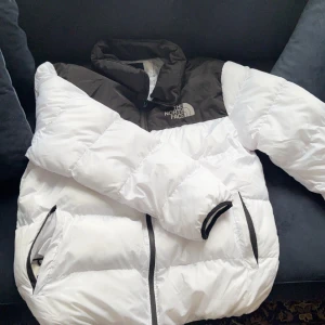 Vit och svart dunjacka från The North Face - Säljer en snygg vit och svart dunjacka från The North Face. Jackan har en pufferdesign med dragkedja och hög krage för extra värme. Perfekt för kyliga dagar.