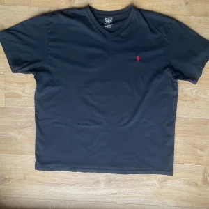 Svart t-shirt från Ralph Lauren - Säljer en klassisk svart t-shirt från Ralph Lauren med en röd broderad logotyp på bröstet. T-shirten har en v-ringad hals och korta ärmar. Perfekt för en stilren look.