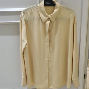 Skjorta - Snygg beige skjorta från NA-KD med klassisk krage och knappar framtill. Perfekt för en stilren look. Skjortan har långa ärmar och en avslappnad passform.