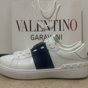 Valentino Garavani sneakers - Snygga vita sneakers från Valentino Garavani med en bred blå rand på sidan. Skorna har en klassisk snörning och en unik design med nitar på sulan. Perfekta för en stilren look.