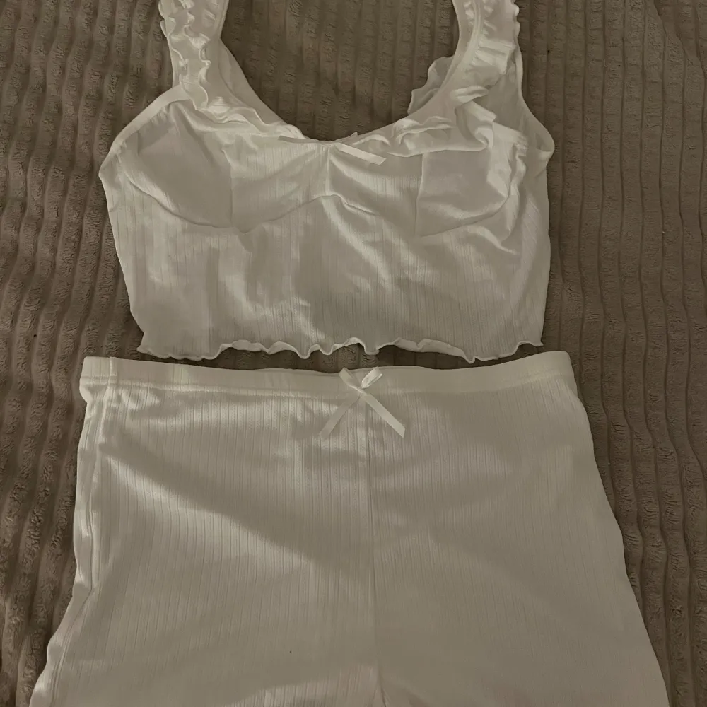 Säljer en söt vit ribbad linnetopp och shorts med volangdetaljer vid axelbanden. Toppen är croppad och har en liten rosett framtill. Perfekt för varma sommardagar!. Yläosat.