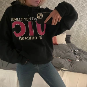 Svart hoodie med UIC-tryck - Säljer en svart hoodie med stort UIC-tryck i vitt och rosa på framsidan. Hoodien har en klassisk passform med en stor ficka framtill och justerbar huva. Perfekt för en avslappnad stil.