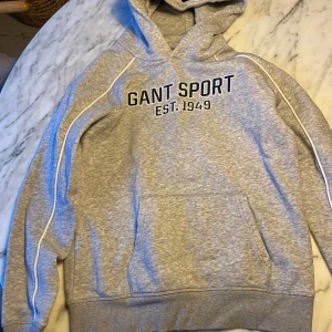 Grå hoodie från GANT - Säljer en snygg grå hoodie från GANT med texten 'GANT SPORT EST. 1949' på framsidan. Tröjan har en klassisk känguruficka och en bekväm huva. Perfekt för en avslappnad stil.