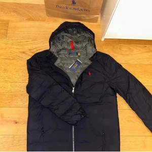 Svart dunjacka från Polo Ralph Lauren - Säljer en stilren svart dunjacka från Polo Ralph Lauren. Jackan har en dragkedja framtill och är lätt att packa ihop i en liten väska. Den är perfekt för kyligare dagar. Tagen tillkommer inte