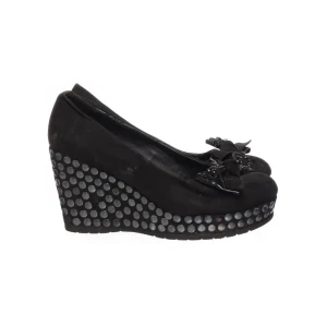 Svarta kilklackar med rosett - Snygga svarta kilklackar med dekorativa rosetter och nitar på sulan.Några av nitarna är borta och dom är ganska slitna men de funkar fof. Skorna har en hög kilklack och en rundad tå.