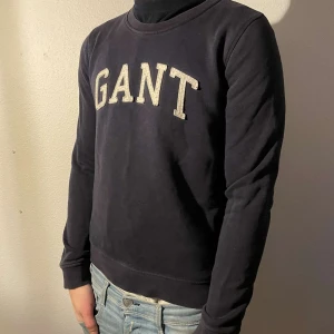 Svart sweatshirt från GANT - Säljer en stilren svart sweatshirt från GANT med deras logga i vitt på bröstet. Tröjan har långa ärmar och en rund halsringning. Perfekt för en avslappnad stil.