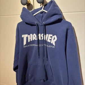 Blå hoodie från Thrasher - Säljer en blå hoodie från Thrasher med vit text på bröstet. Hoodien har en liten fläck som inte går bort och sen liten rivning men annars är den i bra skick och inga andra skador på den. Den är i storlek S.