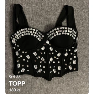 Svart korsett med strassdetaljer  - Snygg svart korsett med glittrande strassdetaljer och dragkedja bak. Perfekt för att ge en extra touch till din outfit. Den har breda axelband och en figurnära passform.