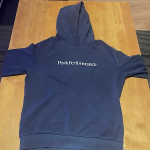 Mörkblå hoodie från Peak Performance - Säljer en snygg mörkblå hoodie från Peak Performance med vit logga på bröstet. Tröjan har en klassisk känguruficka och en bekväm huva. Perfekt för kyliga dagar!