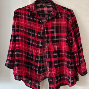 Röd och svart rutig skjorta - Snygg röd och svart rutig skjorta med knappar framtill. Skjortan har en avslappnad passform och trekvartsärmar, perfekt för en casual look. Den är tillverkad i ett mjukt material som känns skönt mot huden.