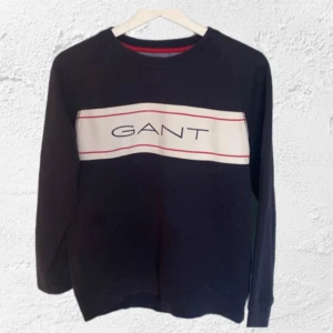  sweatshirt  GANT - Säljer en stilren mörkblå sweatshirt från GANT med vit och röd detalj över bröstet och GANT-logga. Tröjan har långa ärmar och en bekväm passform. Perfekt för en avslappnad stil.