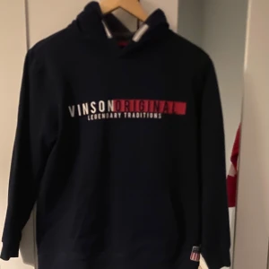 Svart hoodie från Vinson - Säljer en svart hoodie från Vinson med texten 'Vinson Original Legendary Traditions' i vitt och rött på framsidan. Tröjan har en klassisk design med långa ärmar och en bekväm huva. Perfekt för en avslappnad stil.