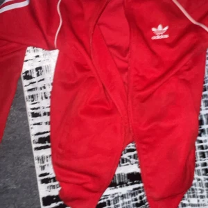 Röd träningsjacka från Adidas - Säljer en röd träningsjacka från Adidas med vita detaljer och logga på bröstet. Jackan har långa ärmar och dragkedja framtill. Perfekt för sportiga aktiviteter eller en avslappnad stil.